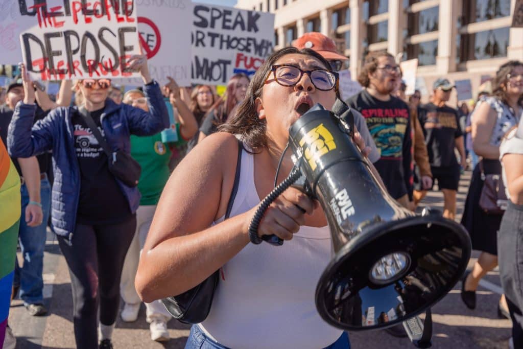 Una manifestante encabeza la protesta con megáfono en mano, mientras decenas de personas avanzan detrás con carteles y consignas. La movilización refleja el rechazo a políticas migratorias y la organización comunitaria en el espacio público.